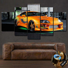 Load image into Gallery viewer, Multi-panel canvas wall art featuring a bright orange Toyota Supra sports car in an industrial setting above a brown leather sofa