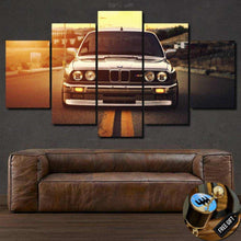 Load image into Gallery viewer, BMW E30 M3 Canvas wall art featuring a front view of the car in sunset tones above a brown leather sofa, free shipping worldwide